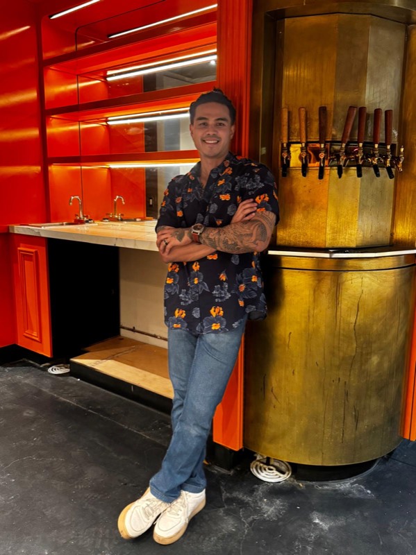Ethan, co-founder of Wanjai, smiling with arms crossed beside the brass beer taps and the orange back bar