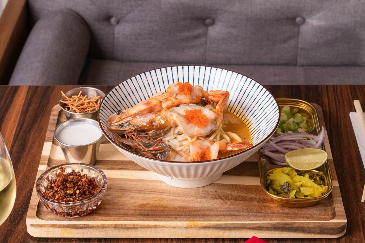 Khao soi topped with whole prawns and orange roe in a striped bowl, served on a wood board with crispy noodles, coconut milk, chili, red onion, lime, and pickled greens