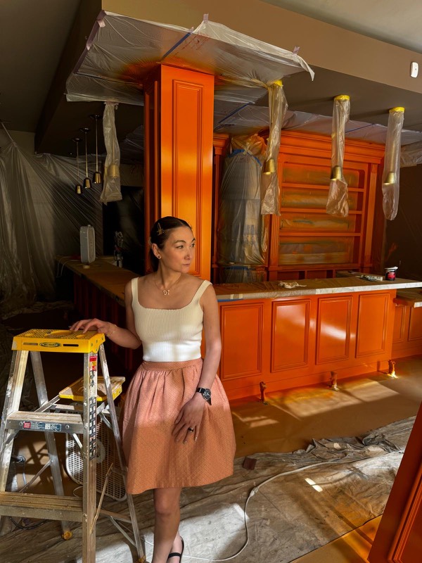 Kitty, co-founder of Wanjai, leaning on a yellow ladder in front of the freshly painted orange bar, with the lights still wrapped in plastic