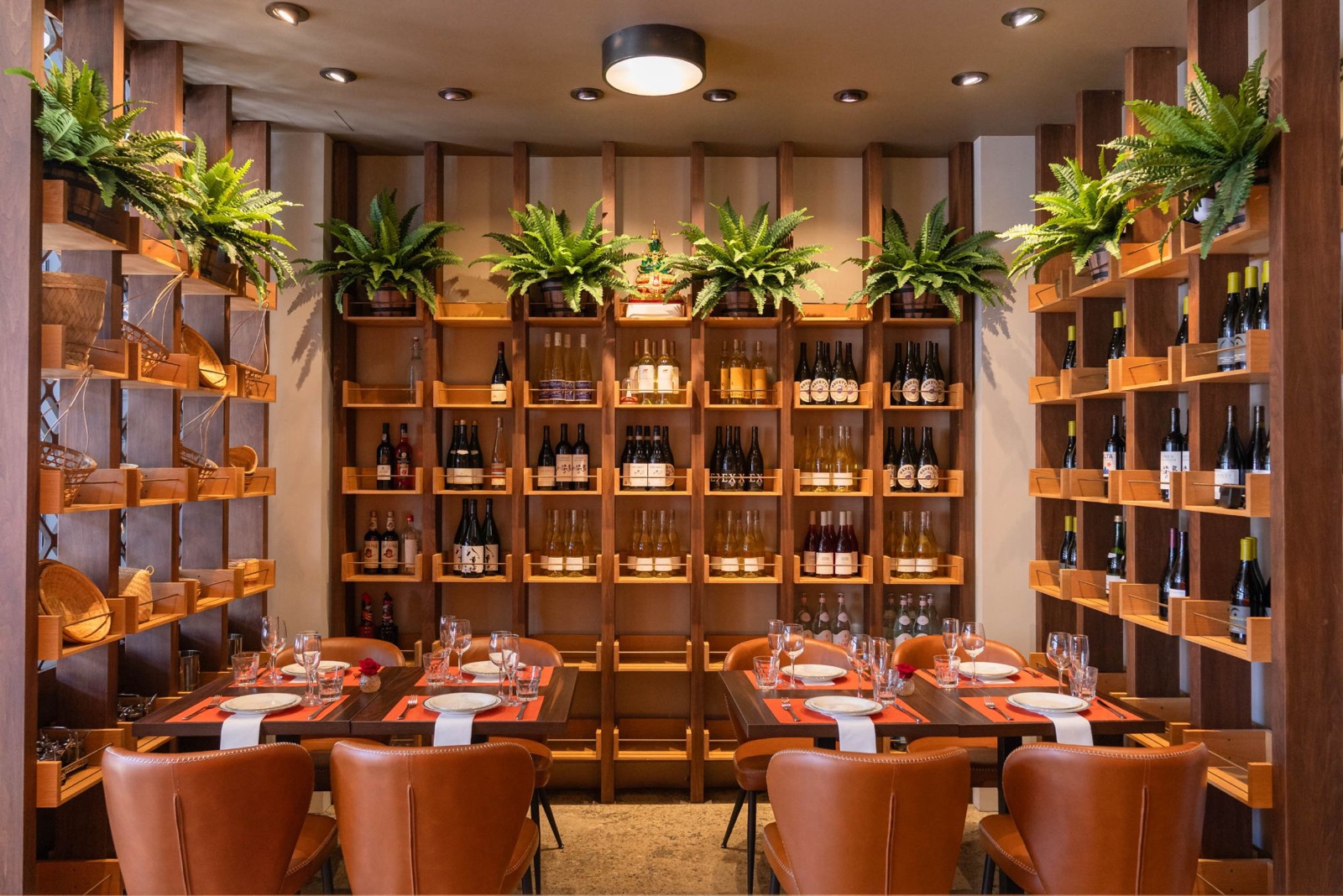 Two tables set with orange placemats and wine glasses in front of Wanjai's wall of wine shelves, lined with ferns and woven baskets