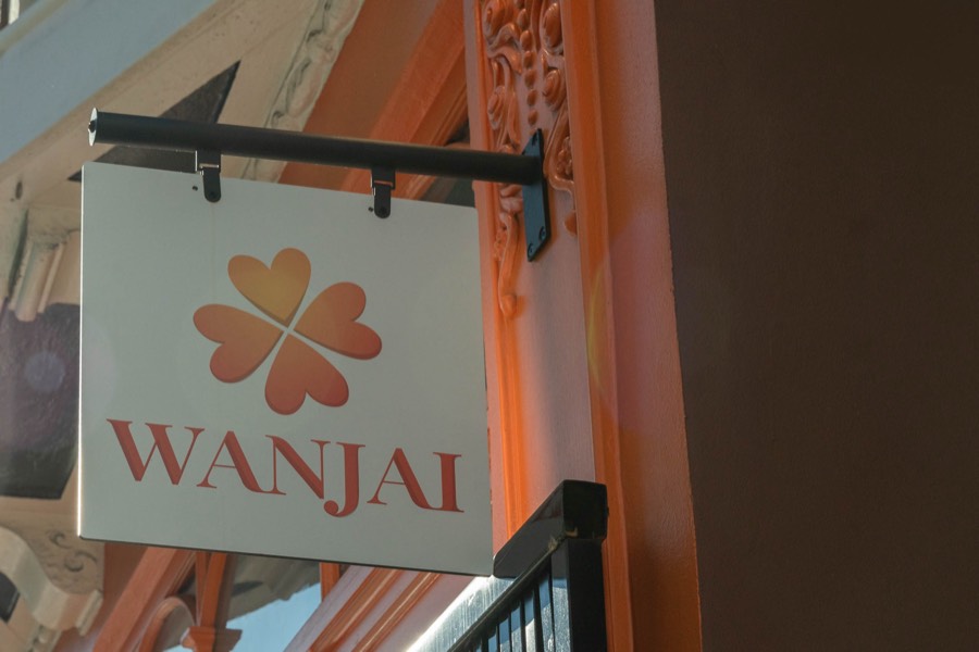 The white Wanjai sign with the orange clover logo, hanging from a black bracket beside the orange facade