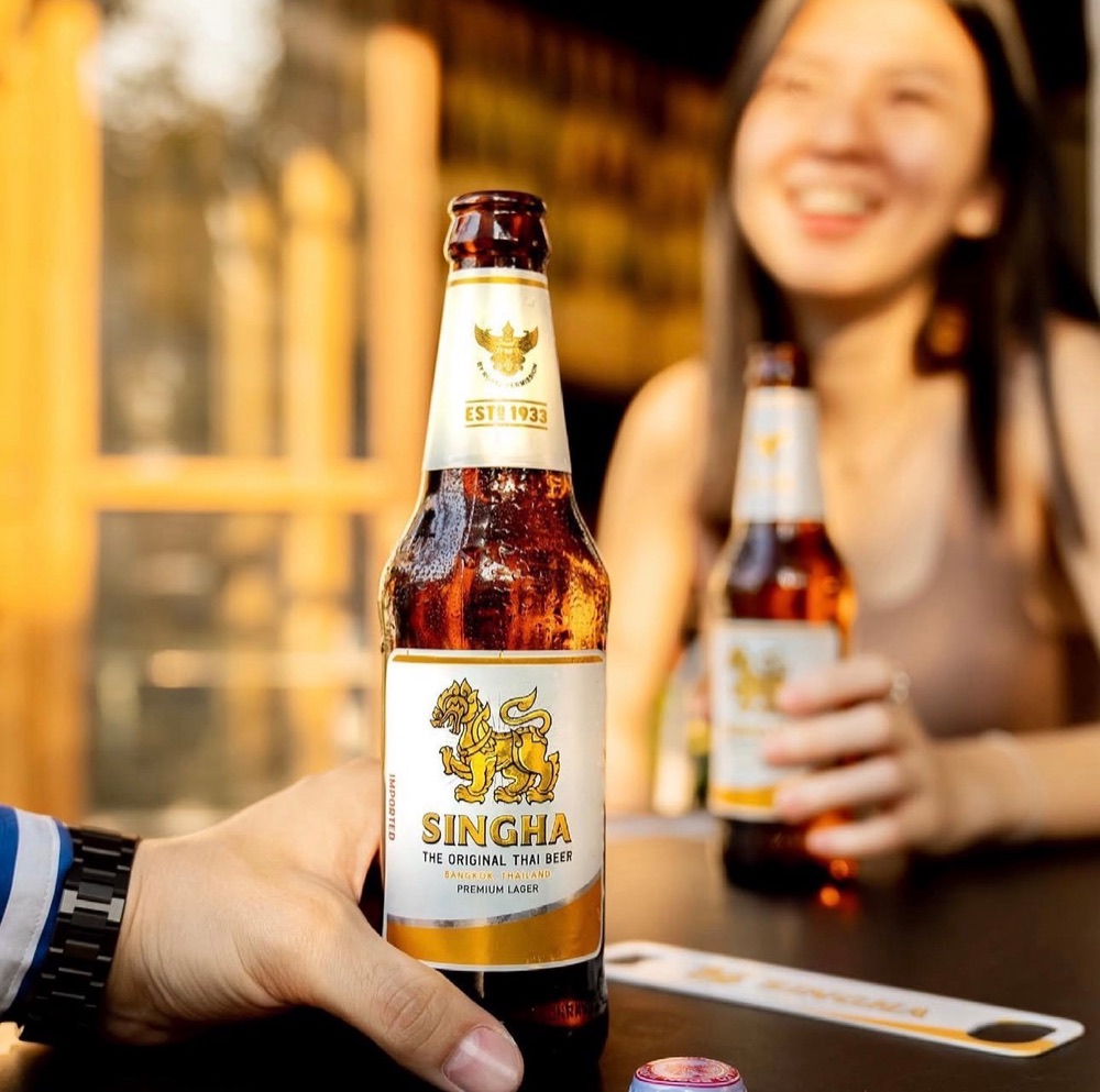 A cold bottle of Singha beer held up at the table, with a smiling friend holding another behind it